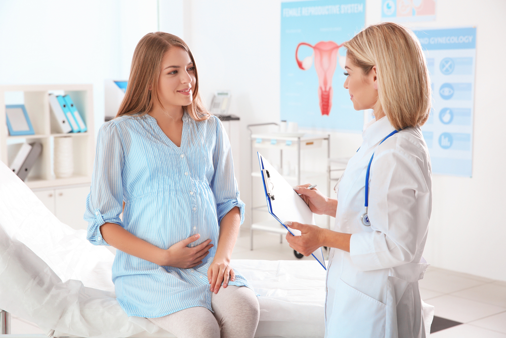 Pregnant woman sitting on doctor's table talking to female doctor holding clipboard.